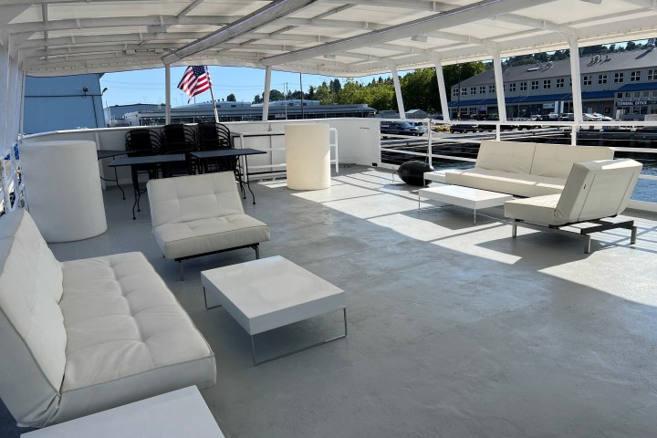 Open boat deck with white sofas, tables, chairs and an American flag under a canopy.