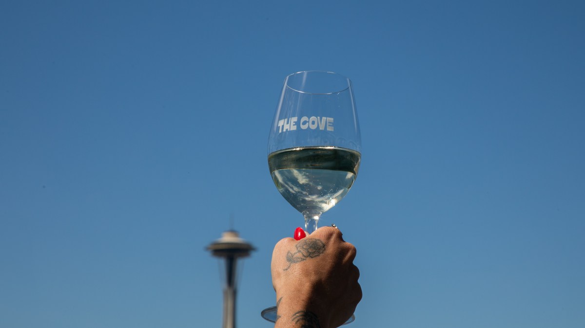 Hand holding a wine glass reading 'The Cove' with Space Needle in background.