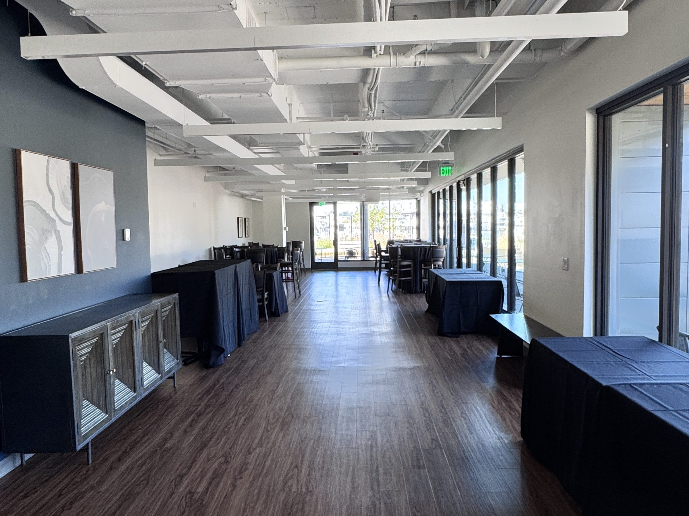 Long hallway with dark wooden floor, tables with black cloth, and large windows on the right.