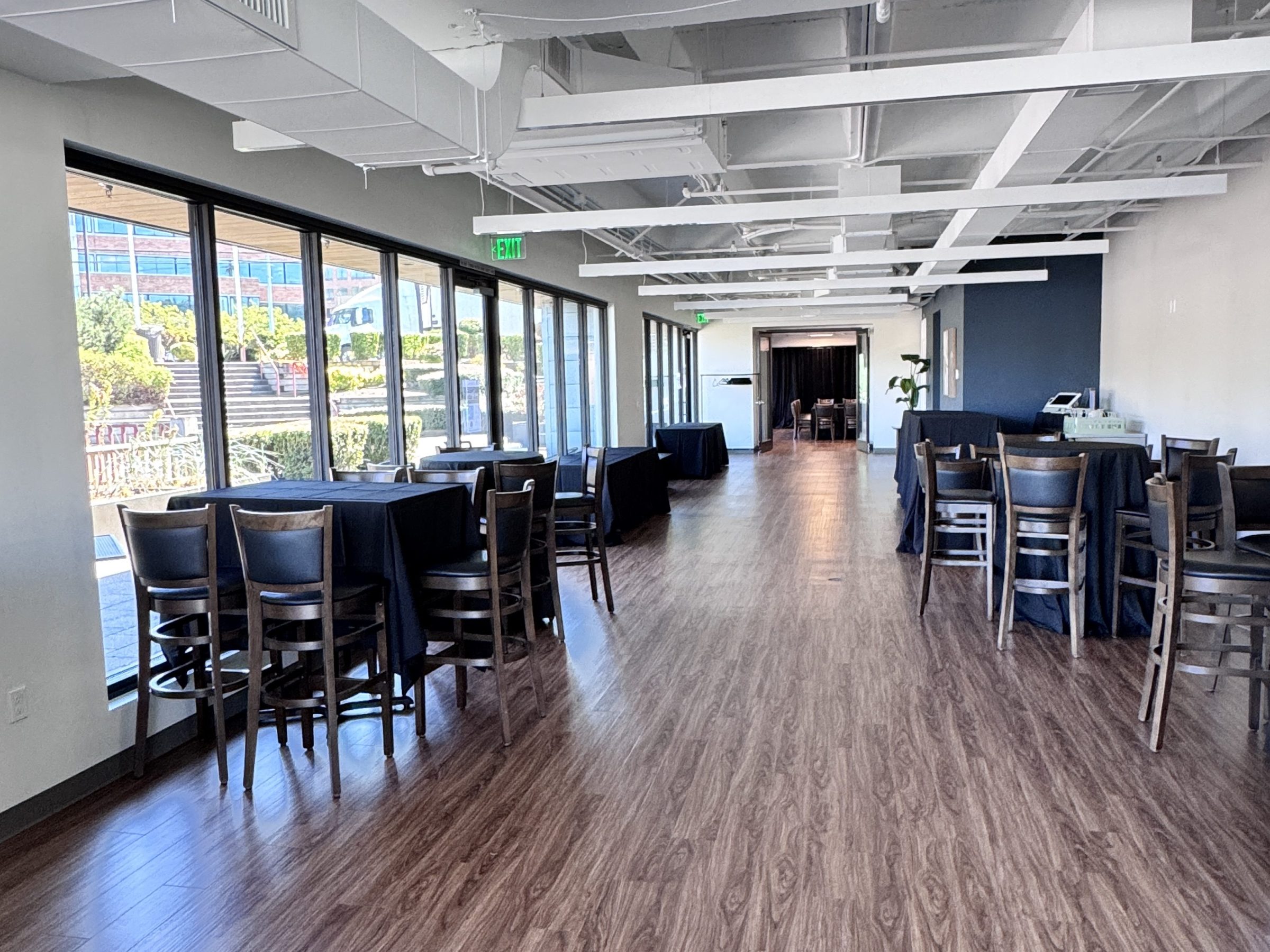Bright room with wooden floor, large windows, tables, and chairs, set up for an event.