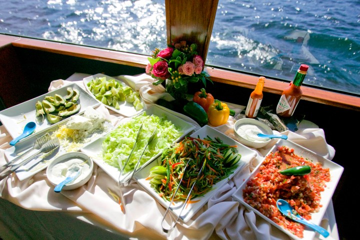 Buffet with salads, avocado, sauces, and condiments on a table by a sunlit window with ocean view.
