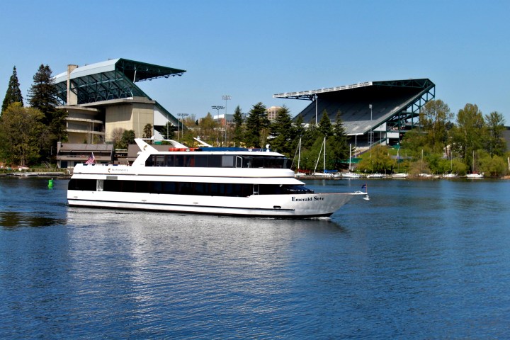 White yacht on a lake near a large stadium with trees in background.