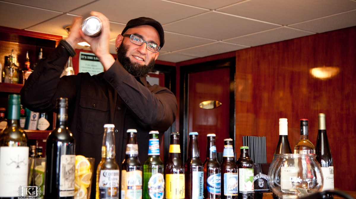 Bartender shaking cocktail mixer behind bar with assorted bottles.