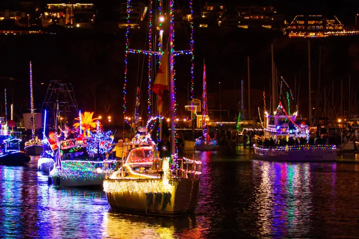 Boats adorned with colorful holiday lights reflecting on water at night.